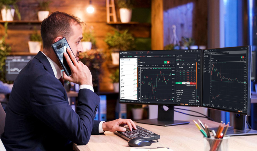 Stock broker analyzing live market charts on multiple screens while assisting a client over a phone call.
